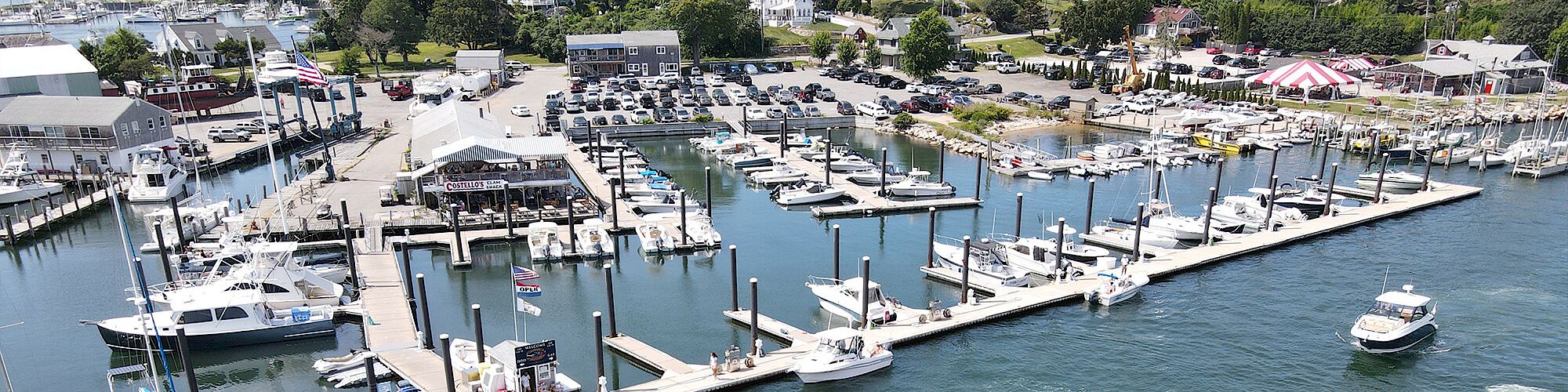 The image depicts a marina with several boats docked at piers and a few boats navigating on the water. The surrounding area includes buildings and trees.