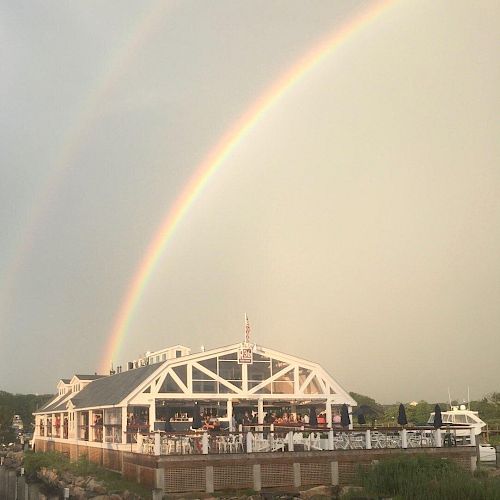 The image shows a waterfront building under a double rainbow with a cloudy sky in the background. The building appears to be a restaurant or caf&eacute;.