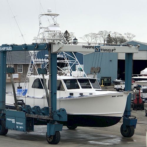 A white boat suspended by a large blue Marine Travelift with "Noank Shipyard" written on it, in an industrial marina area with other boats and buildings.